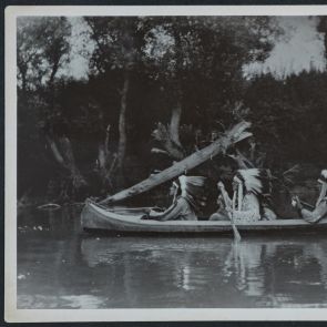 Three Indians with headdresses paddle in canoe